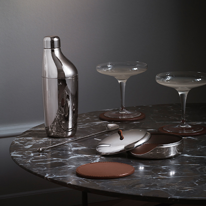 bar shaker and coasters mirror polished stainless steel and brown leatcher on a dark coloured marble with black background from Sky collection by Georg Jensen