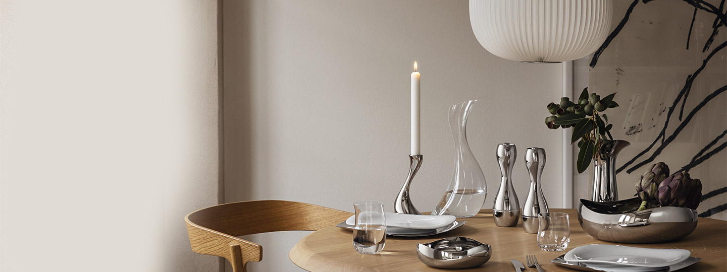 salt and pepper grinder, small and large bowl, candle holder in mirror polished stainless steel in a dinner setting on a brown table from the cobra collection by georg jensen