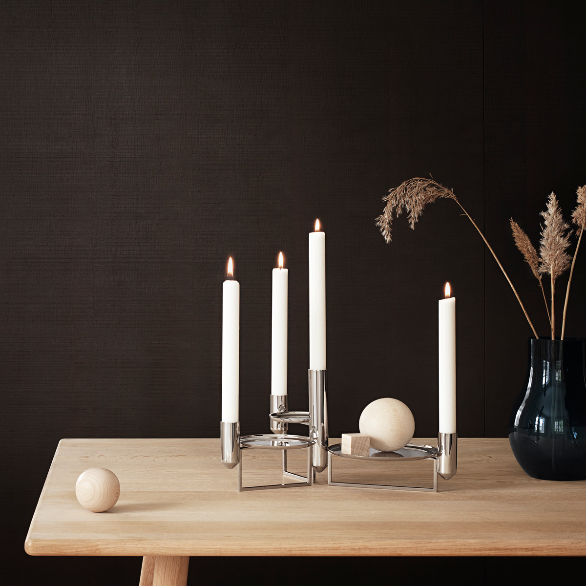 Set of three white taper candles in a chrome candle holder on a wooden table with a dark background and neutral decor.